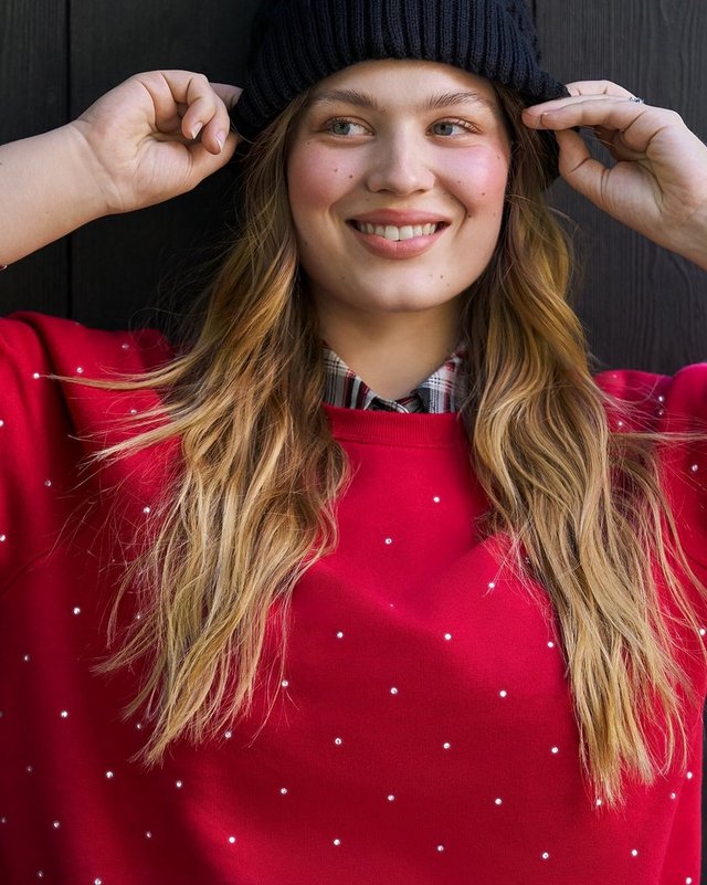 A model with long blonde hair smiles at the camera against a dark vertical wood plank wall, both hands raised to the brim of a navy ribbed knit beanie. She wears a red sweatshirt scattered with small white dots, with a plaid collared shirt layered underneath and its collar showing at the neckline.