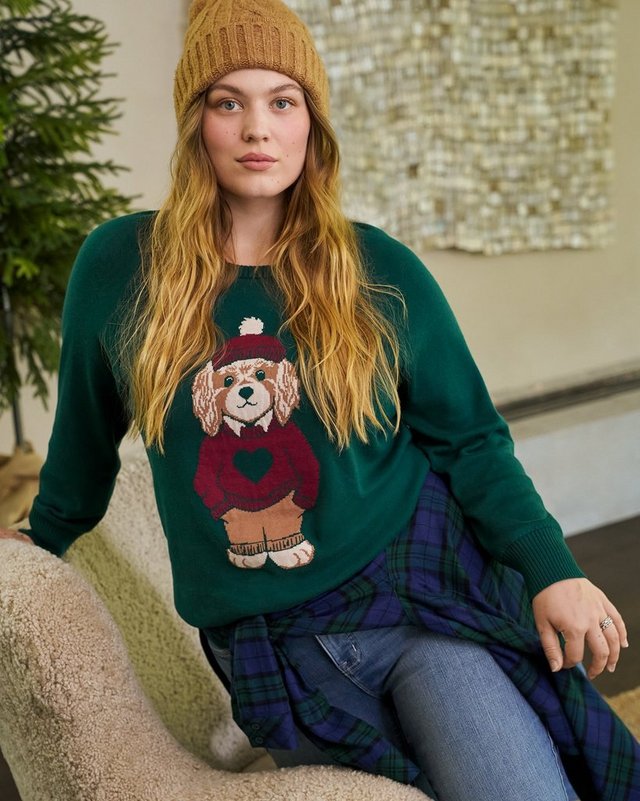 A model with long strawberry blonde hair sits leaning against a pale surface in front of a softly lit background, smiling at the camera. She wears a camel ribbed knit beanie and a dark green sweatshirt printed with a cartoon dog in a red sweater and scarf. A navy and green plaid skirt or pants shows at the bottom of the frame.