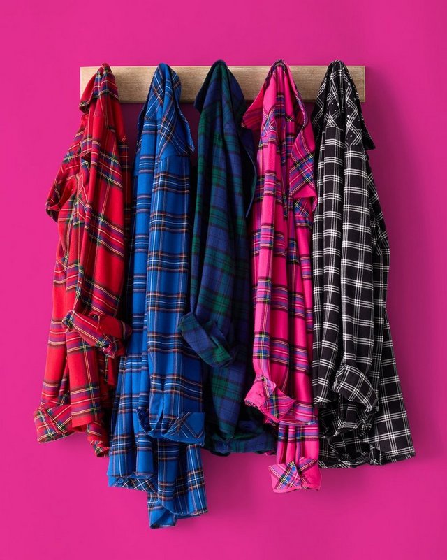 Five plaid flannel shirts hang side by side on black hangers from a wooden rail mounted against a bright magenta pink background. From left to right, the shirts are red and black tartan, bright blue and black plaid, dark green and navy plaid, hot pink and black plaid, and black and white check. The shirts hang open and overlap slightly, their tails falling at uneven lengths.