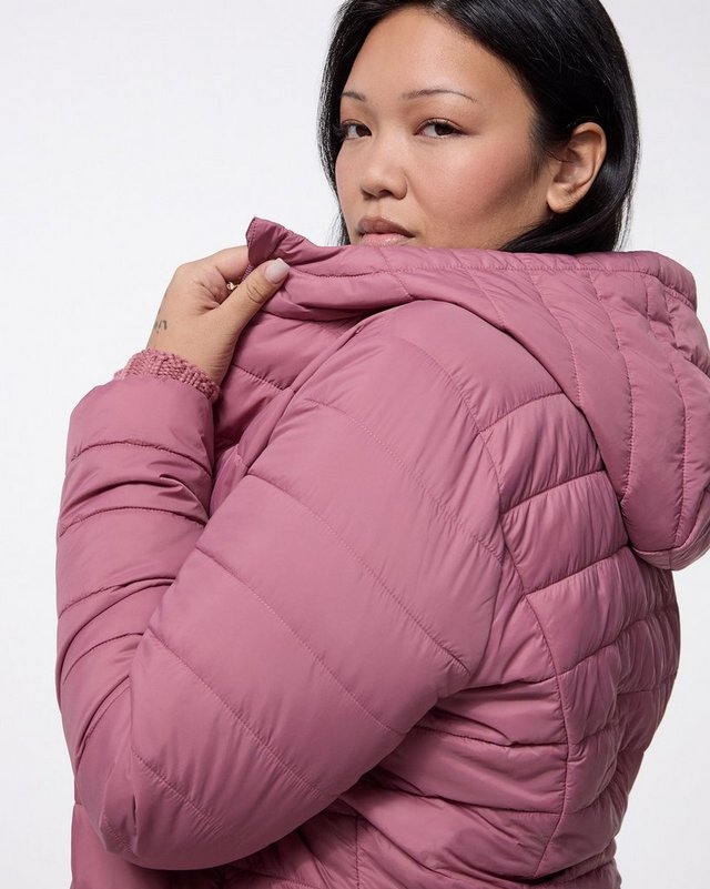 A close-up of a model with dark hair pulled back, shown from the head to the waist against a plain pale grey background, her head turned to look toward the camera and one hand raised to the collar of her jacket. She wears a dusty rose puffer jacket with horizontal quilted channels, a padded hood, long sleeves with elasticated cuffs, and a raised collar. A mauve ribbed cuff shows at her wrist.