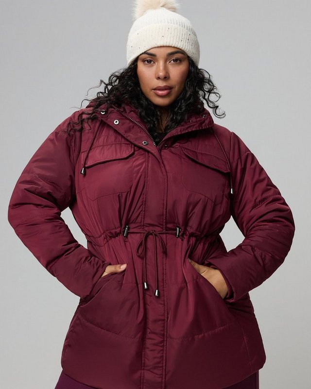 A model with dark curly hair stands facing the camera against a plain pale grey background, both hands tucked into her coat pockets. She wears a cream ribbed knit beanie with a folded brim and a long burgundy puffer coat with a stand collar, a snap-front storm placket over a zip, a hood, chest flap pockets, side slash pockets, and a drawstring cinched at the waist with cord ends hanging down. The quilted panels give a padded horizontal texture through the body.