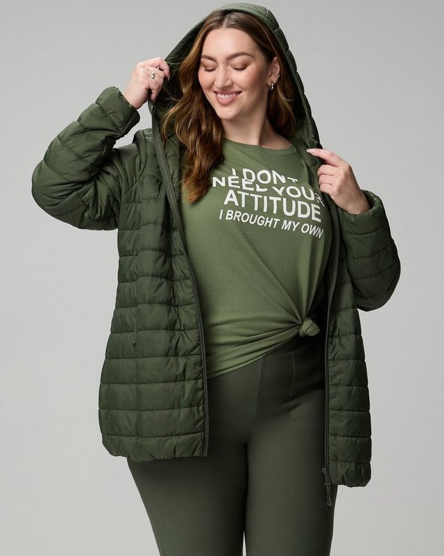 A model with long auburn hair stands against a plain white background, smiling with her head tilted down and one hand raised to hold the edge of her hood. She wears an olive green quilted puffer jacket worn open, with a padded hood, a full zip front, and vertical quilted channels. Underneath is a light olive green T-shirt knotted at the front, printed in white type reading ‘DON’T NEED YOUR ATTITUDE’ above ‘I BROUGHT MY OWN’. She wears matching olive green leggings.