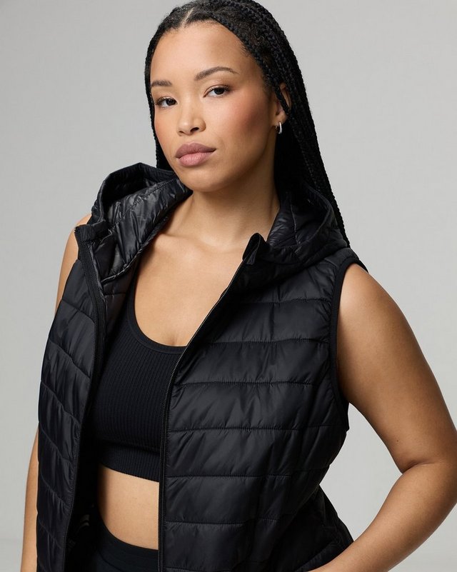 A model with long braided hair is shown from the head to the hips against a plain white background, looking directly at the camera with one hand on her hip. She wears a black quilted puffer vest worn open, with vertical quilted channels, a padded hood, and a zip front. Underneath is a black sports bra with a scoop neckline, leaving her midriff bare, and black bottoms show at the bottom edge. She wears small hoop earrings.