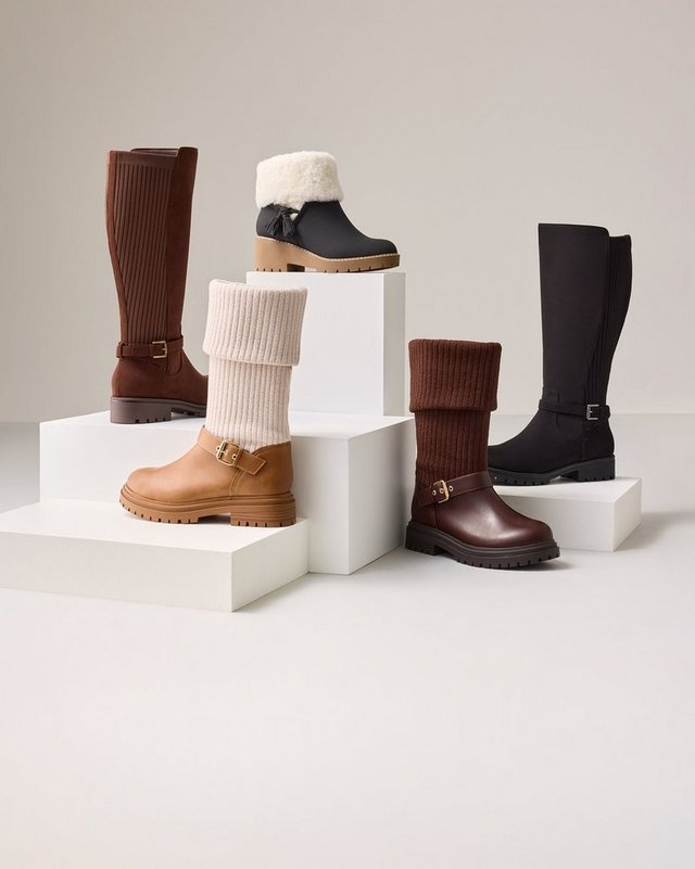 Five boots arranged on white blocks of varying heights against a plain white background. At the back left stands a tall cognac brown knee-high boot with a buckled ankle strap and a low heel. Behind it on a raised block is a short dark brown boot with a cream shearling collar and a lugged sole. In the center front is a tan ankle boot with a thick ribbed cream knit cuff folded over the shaft and a chunky treaded sole. To its right sits a dark brown boot with a brown ribbed knit cuff, a buckled strap at the ankle, and a lugged sole. At the back right stands a tall black knee-high boot with a smooth shaft, a buckled ankle strap, and a flat sole.