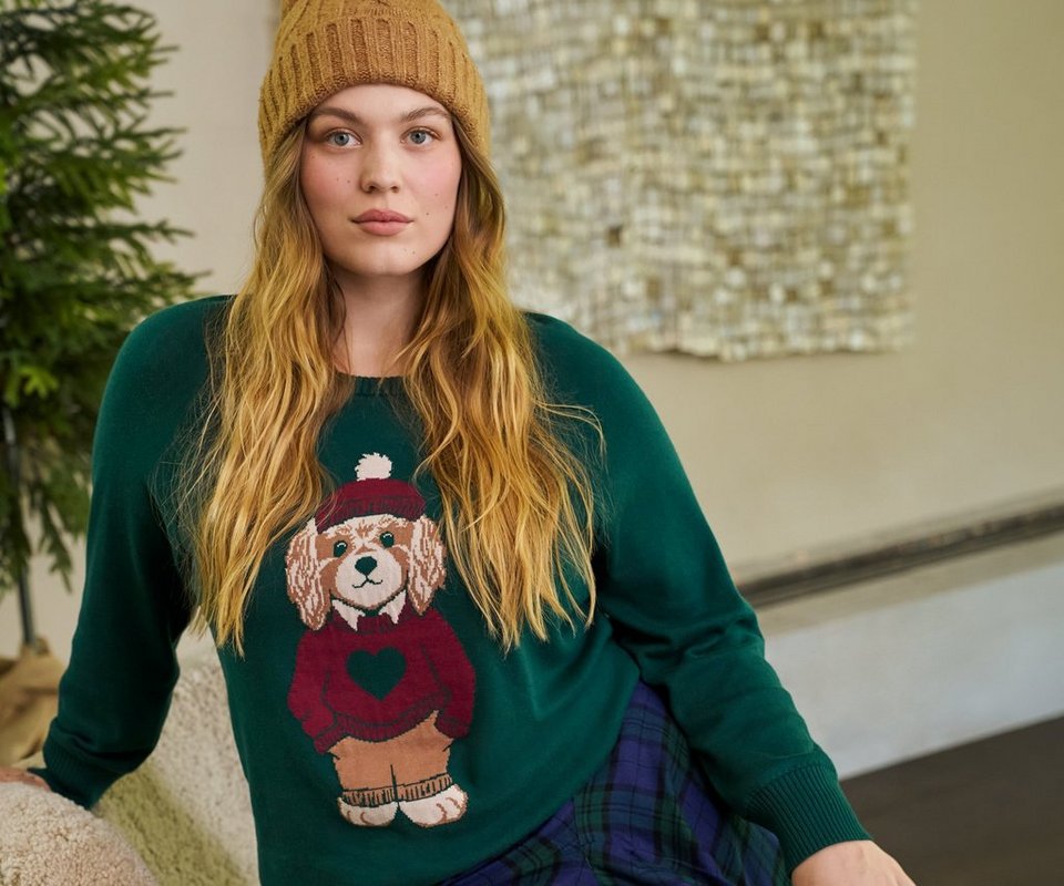 A model with long strawberry blonde hair sits leaning against a pale surface in front of a softly lit background, smiling at the camera. She wears a camel ribbed knit beanie and a dark green sweatshirt printed with a cartoon dog in a red sweater and scarf. A navy and green plaid skirt or pants shows at the bottom of the frame.