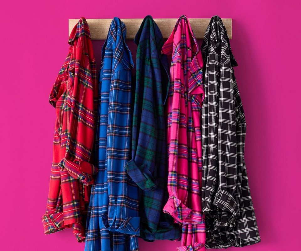 Five plaid flannel shirts hang side by side on black hangers from a wooden rail mounted against a bright magenta pink background. From left to right, the shirts are red and black tartan, bright blue and black plaid, dark green and navy plaid, hot pink and black plaid, and black and white check. The shirts hang open and overlap slightly, their tails falling at uneven lengths.