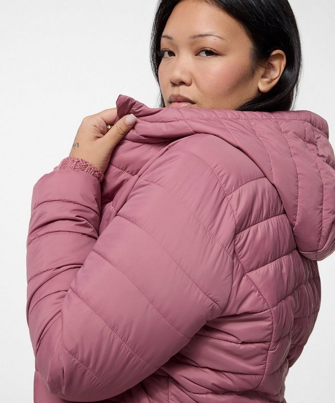 A close-up of a model with dark hair pulled back, shown from the head to the waist against a plain pale grey background, her head turned to look toward the camera and one hand raised to the collar of her jacket. She wears a dusty rose puffer jacket with horizontal quilted channels, a padded hood, long sleeves with elasticated cuffs, and a raised collar. A mauve ribbed cuff shows at her wrist.