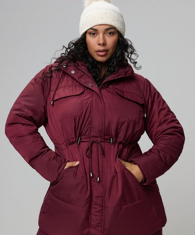A model with dark curly hair stands facing the camera against a plain pale grey background, both hands tucked into her coat pockets. She wears a cream ribbed knit beanie with a folded brim and a long burgundy puffer coat with a stand collar, a snap-front storm placket over a zip, a hood, chest flap pockets, side slash pockets, and a drawstring cinched at the waist with cord ends hanging down. The quilted panels give a padded horizontal texture through the body.