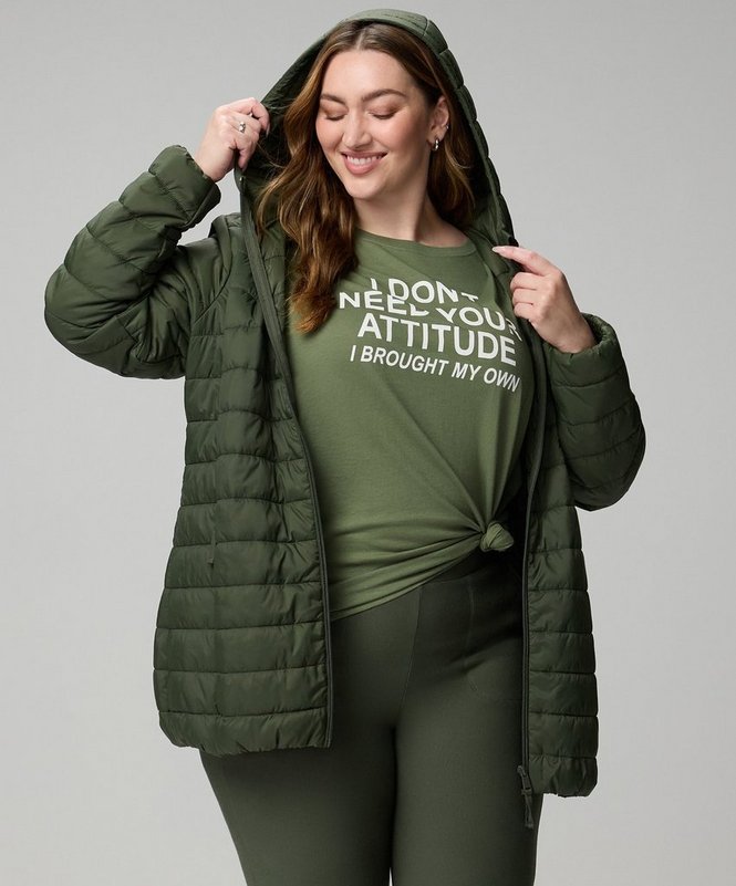A model with long auburn hair stands against a plain white background, smiling with her head tilted down and one hand raised to hold the edge of her hood. She wears an olive green quilted puffer jacket worn open, with a padded hood, a full zip front, and vertical quilted channels. Underneath is a light olive green T-shirt knotted at the front, printed in white type reading ‘DON’T NEED YOUR ATTITUDE’ above ‘I BROUGHT MY OWN’. She wears matching olive green leggings.