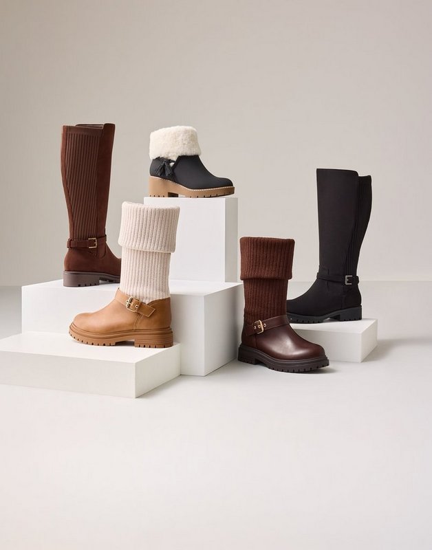 Five boots arranged on white blocks of varying heights against a plain white background. At the back left stands a tall cognac brown knee-high boot with a buckled ankle strap and a low heel. Behind it on a raised block is a short dark brown boot with a cream shearling collar and a lugged sole. In the center front is a tan ankle boot with a thick ribbed cream knit cuff folded over the shaft and a chunky treaded sole. To its right sits a dark brown boot with a brown ribbed knit cuff, a buckled strap at the ankle, and a lugged sole. At the back right stands a tall black knee-high boot with a smooth shaft, a buckled ankle strap, and a flat sole.