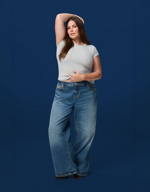 A model with long brown hair stands against a solid dark navy background, one arm raised and bent over her head with the other hand resting on her stomach. She wears a fitted pale grey short-sleeve ribbed tee tucked in, and light-wash blue jeans with a very wide, exaggerated leg that falls full and loose to the floor, with fading down the front of the thighs and small embellishments at the front pocket. The hems pool over black shoes just visible at the bottom.