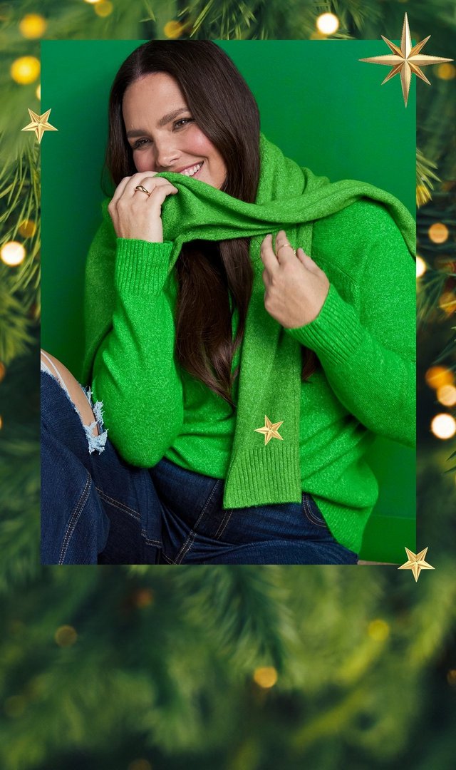 A single panel framed by blurred pine branches, warm golden bokeh lights, and scattered gold star shapes. A model with long brown hair sits against a bright green background, smiling as she holds a green knit scarf up across her chin with both hands. She wears a matching bright green knit sweater with ribbed cuffs, dark-wash jeans, and a ring on one hand.