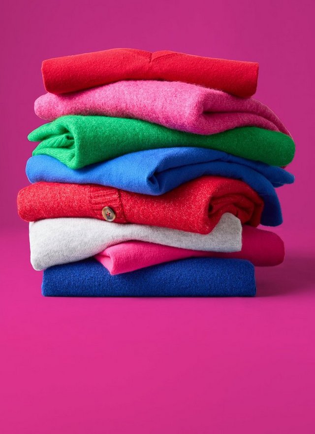 A stack of folded knit sweaters against a bright magenta pink background. From top to bottom the colors run red, pink, green, blue, red with a visible button, cream, pink, and navy, with the folded sleeves and edges creating uneven layered lines across the stack.l’s shoulder.