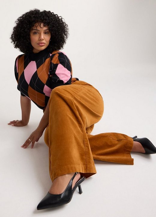 A model with voluminous dark curly hair kneels and leans forward on a plain white studio floor, one hand planted on the ground and her legs folded to one side. She wears a short-sleeve knit top in a large argyle pattern of black, pink, and rust brown diamonds with fine white crosshatch lines, finished with a ribbed mock neck and ribbed cuffs. Her pants are tan corduroy with a wide leg, and she wears black pointed-toe slingback heels with a stiletto heel. She wears a ring on each hand.