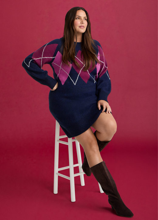 A model with long brown hair sits on a white wooden stool against a deep raspberry red background, one hand on her hip and one leg crossed over the other. She wears a navy knit sweater dress with a large argyle pattern across the chest and sleeves in magenta, pink, and white diamond outlines, with a crewneck, long blouson sleeves, ribbed cuffs, and a ribbed hem ending above the knee. On her feet are dark brown suede knee-high boots.