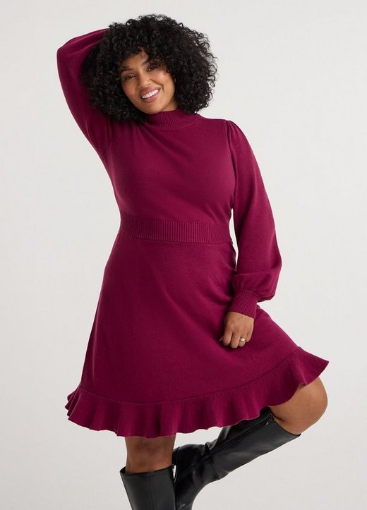 A model with dark curly hair smiles with one arm raised over her head against a plain white background. She wears a deep magenta knit dress with a ribbed mock neck, long sleeves gathered at the shoulder with ribbed cuffs, a wide ribbed waistband, and a flared skirt finished with a pleated ruffle hem above the knee. On her feet are black knee-high boots, and she wears a ring on one hand.