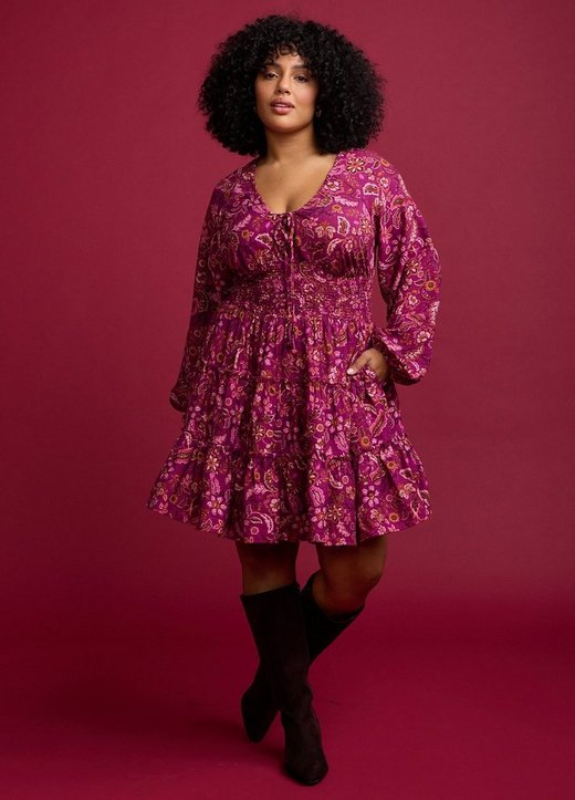 A model with voluminous dark curly hair stands against a deep raspberry red background, one leg crossed behind the other and one hand in her pocket. She wears a magenta mini dress printed with an allover paisley and floral pattern in pink, rust, and cream, with a V neckline, a drawstring tie at the chest, long blouson sleeves with elasticated cuffs, a smocked waist panel, and a tiered ruffled skirt. On her feet are dark brown suede knee-high boots, and she wears small gold hoop earrings.