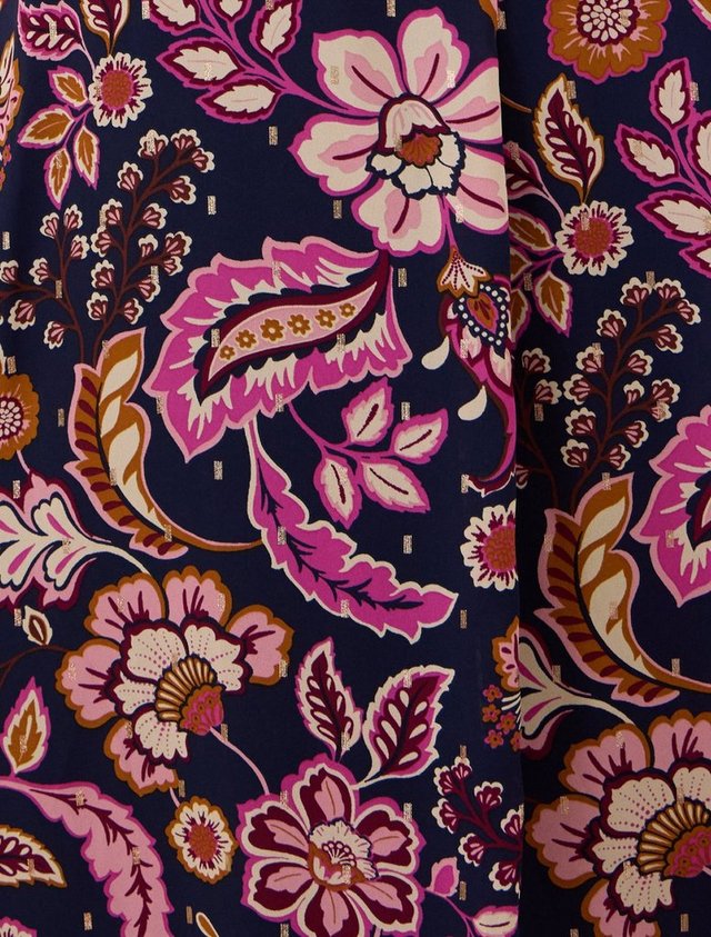 An extreme close-up of printed fabric filling the frame, folded once so a horizontal crease runs across the middle. The navy ground is covered in a large-scale botanical print of oversized blooms, curling leaves, and paisley forms in bright pink, magenta, pale pink, cream, burgundy, and golden ochre. Small metallic gold dashes are scattered across the open navy areas between the motifs. There are no people, objects, or text in the image.