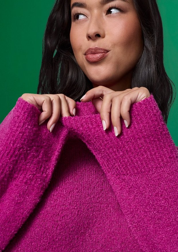 A close-up of a model with long dark hair against a green background, her head turned to look off to her left with her hands holding the folded collar of her sweater. She wears a bright magenta pink sweater in a soft textured knit with a wide ribbed turtleneck collar folded down and ribbed cuffs. Her nails are manicured in a pale shade.