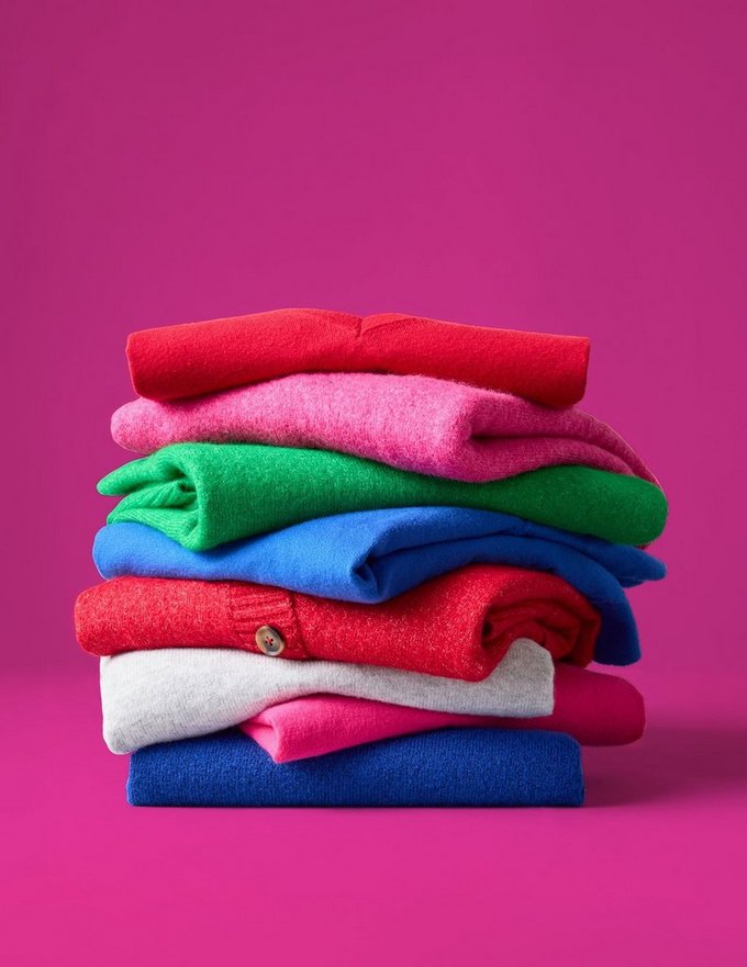 A stack of folded knit sweaters against a bright magenta pink background. From top to bottom the colors run red, pink, green, blue, red with a visible button, cream, pink, and navy, with the folded sleeves and edges creating uneven layered lines across the stack.