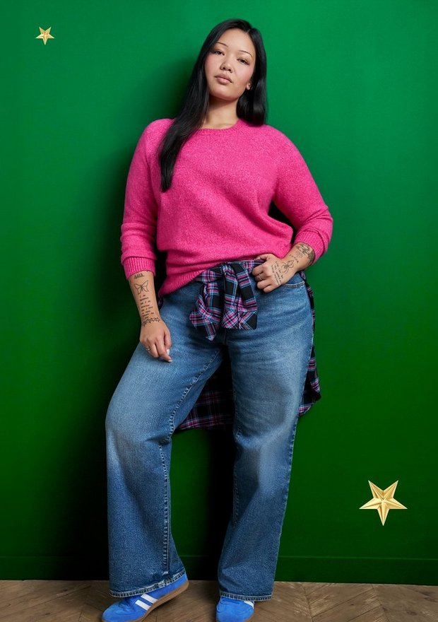 A model with long black hair stands against a green background scattered with gold star shapes, looking at the camera with her arms at her sides. She wears a hot pink knit crewneck sweater with a dark plaid flannel shirt tied around her waist, and light-wash wide-leg jeans. Her shoes are blue sneakers, and tattoos are visible on both forearms.