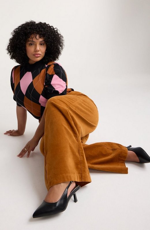 A model with voluminous dark curly hair kneels and leans forward on a plain white studio floor, one hand planted on the ground and her legs folded to one side. She wears a short-sleeve knit top in a large argyle pattern of black, pink, and rust brown diamonds with fine white crosshatch lines, finished with a ribbed mock neck and ribbed cuffs. Her pants are tan corduroy with a wide leg, and she wears black pointed-toe slingback heels with a stiletto heel. She wears a ring on each hand.