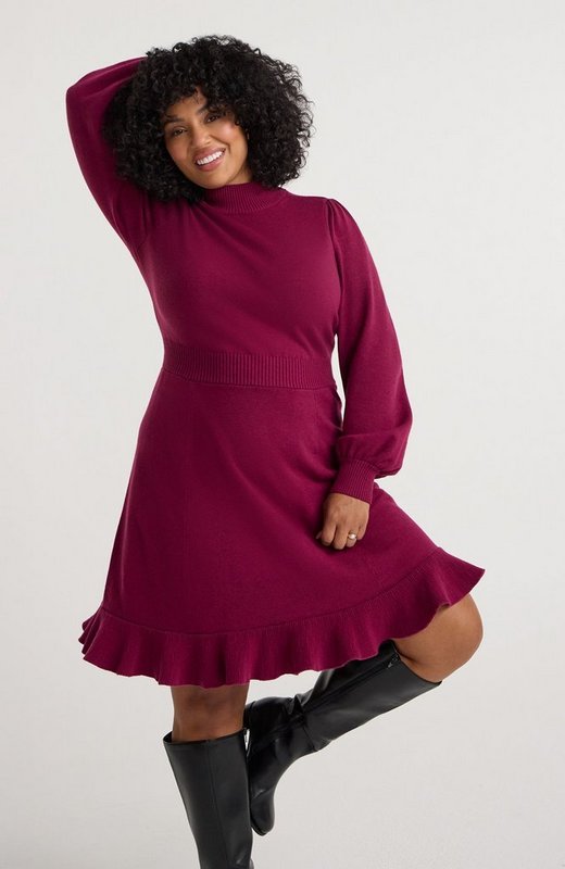 A model with dark curly hair smiles with one arm raised over her head against a plain white background. She wears a deep magenta knit dress with a ribbed mock neck, long sleeves gathered at the shoulder with ribbed cuffs, a wide ribbed waistband, and a flared skirt finished with a pleated ruffle hem above the knee. On her feet are black knee-high boots, and she wears a ring on one hand.
