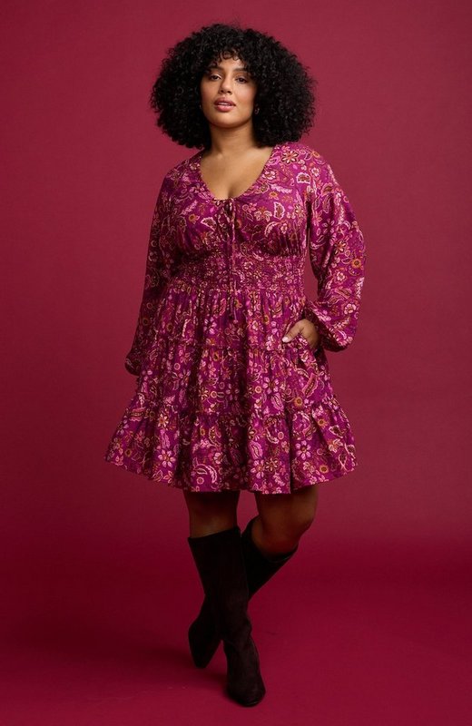 A model with voluminous dark curly hair stands against a deep raspberry red background, one leg crossed behind the other and one hand in her pocket. She wears a magenta mini dress printed with an allover paisley and floral pattern in pink, rust, and cream, with a V neckline, a drawstring tie at the chest, long blouson sleeves with elasticated cuffs, a smocked waist panel, and a tiered ruffled skirt. On her feet are dark brown suede knee-high boots, and she wears small gold hoop earrings.