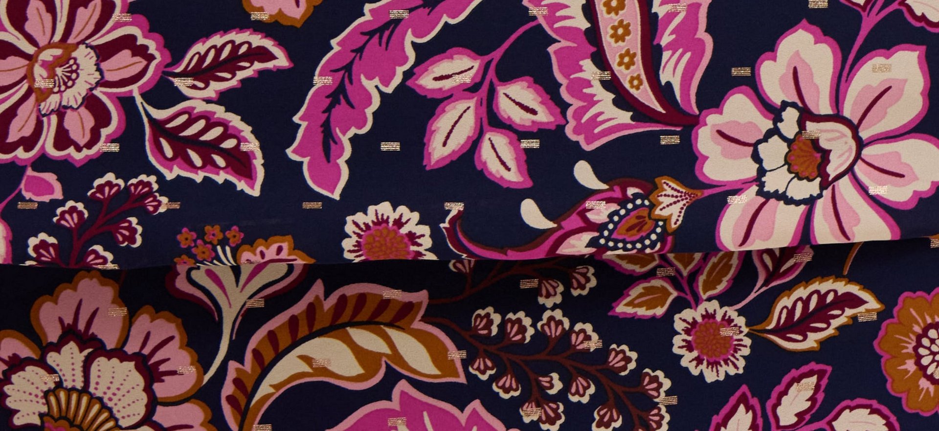 An extreme close-up of printed fabric filling the frame, folded once so a horizontal crease runs across the middle. The navy ground is covered in a large-scale botanical print of oversized blooms, curling leaves, and paisley forms in bright pink, magenta, pale pink, cream, burgundy, and golden ochre. Small metallic gold dashes are scattered across the open navy areas between the motifs. There are no people, objects, or text in the image.
