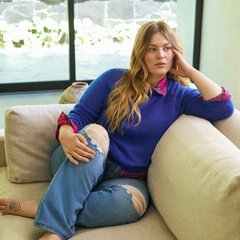 'A model with long wavy strawberry-blonde hair sits on a cream sectional sofa, leaning on the armrest with one hand against her head and her legs drawn up. She wears a cobalt blue crewneck sweater over a pink and purple plaid collared shirt, with the collar and cuffs showing, and light-wash blue jeans with distressed rips at the knees. Behind her, large windows look out onto an outdoor patio with greenery and a stone wall.