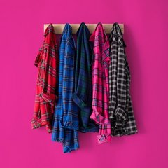 Five plaid flannel shirts hang side by side on black hangers from a wooden rail mounted against a bright magenta pink background. From left to right, the shirts are red and black tartan, bright blue and black plaid, dark navy and blue plaid, hot pink and black plaid, and black and white check. The shirts hang open and overlap slightly, their tails falling at uneven lengths.