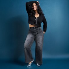 A model with dark curly hair stands against a deep blue background, smiling with one arm raised over her head and her weight shifted to one hip. She wears a cropped black knit cardigan with long sleeves, a V opening, and a silver metallic drawstring tied in a bow at the chest, leaving her midriff bare. Her jeans are washed grey and wide-legged with scattered small embellishments down the front of the legs. On her feet are silver pointed-toe heels.