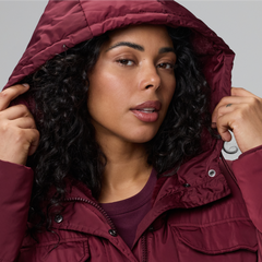 A close-up of a model with long dark curly hair, shown from the head to the upper chest against a plain pale grey background, looking at the camera with both hands raised to the edges of her hood. She wears a burgundy puffer coat with a padded hood, a stand collar, a snap-front placket, and quilted panels. A matching burgundy top shows at the neckline, and she wears a silver watch on her left wrist.