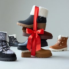 Four winter boots arranged against a pale grey background, with a stack of two in the center tied together by a wide red satin ribbon finished in a large bow. The top boot of the stack is black with a cream shearling cuff and a lugged sole, and below it sits a tan suede boot with a chunky treaded sole. At the left stands a short boot with a grey patterned shaft, white laces, and a black platform sole. At the right stands a tan suede lace-up boot with a cream shearling collar and a lugged sole.