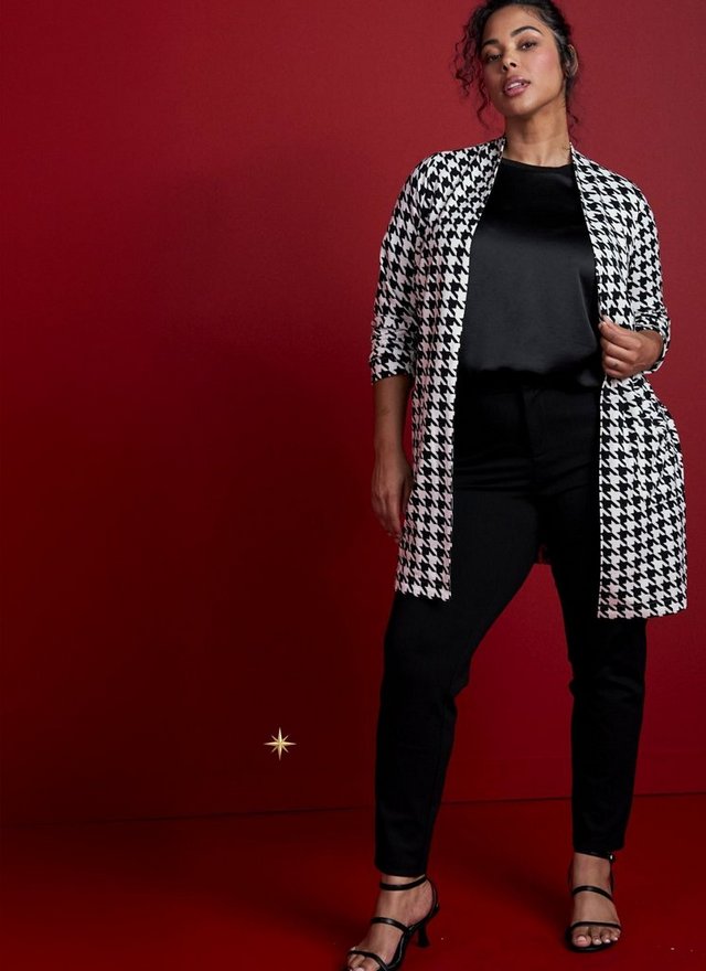 A model with dark curly hair pulled back stands at the right side of a deep red background with a red floor, one hand holding the edge of her cardigan. She wears a long black and white houndstooth duster cardigan worn open, with sleeves pushed up, over a black satin top and black slim-leg pants, with black strappy heeled sandals. A small gold starburst graphic sits on the empty red area at the lower left.