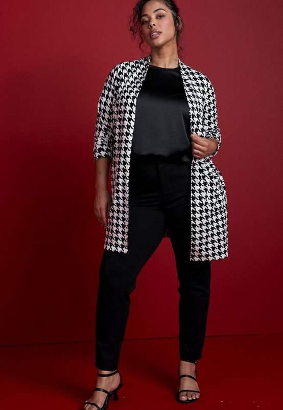 A model with dark curly hair pulled back stands against a deep red background with a red floor, one hand holding the edge of her cardigan. She wears a long black and white houndstooth duster cardigan worn open, with sleeves pushed up the forearm and a straight hem falling below the knee. Underneath is a black satin top and black slim-leg pants. On her feet are black strappy heeled sandals, and she wears a thin gold necklace.