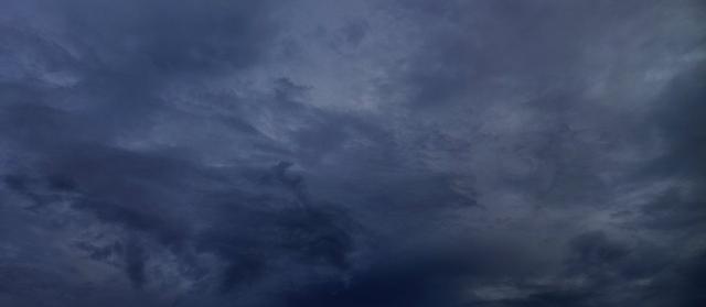A wide, short banner image of an overcast night sky in deep slate blue. Soft layered clouds drift across the frame, lighter and wispier toward the upper right and darker and denser along the lower left, with a small dense cloud formation near the lower center. There are no people, objects, or text in the image.