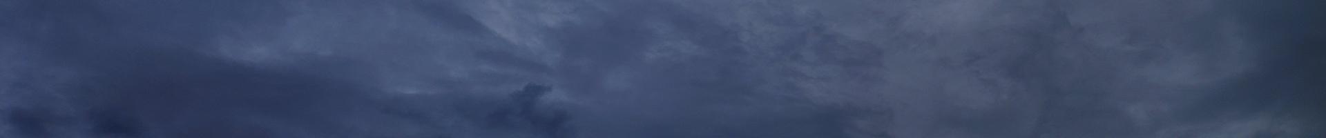 A wide, short banner image of an overcast night sky in deep slate blue. Soft layered clouds drift across the frame, lighter and wispier toward the upper right and darker and denser along the lower left, with a small dense cloud formation near the lower center. There are no people, objects, or text in the image.