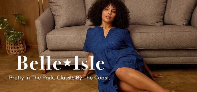 'A wide promotional banner featuring a plus-size model sitting on a herringbone hardwood floor in front of a gray sofa in a warm wood-toned interior. A green leafy plant in a woven basket is visible to the left. The model wears a cobalt blue eyelet short-sleeve midi dress with a v-neckline and lace detail at the bodice, and has voluminous curly dark hair. Overlaid in the lower left in large white serif text reads Belle Isle with a star between the words, and below in smaller white text reads Pretty In The Park. Classic By The Coast.'