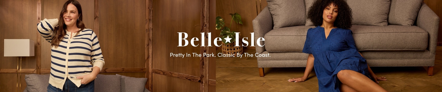 'A wide promotional banner featuring two plus-size models in a warm wood-paneled interior setting with herringbone hardwood floors. On the left, a model stands leaning against a wood-paneled wall beside a gray sofa and a floor lamp, smiling with one hand behind her head. She wears a cream and navy blue horizontal stripe cardigan and dark jeans. In the center, the brand name Belle Isle is displayed in large white serif text with a star between the words, and below in smaller white text reads Pretty In The Park. Classic By The Coast. On the right, a model sits on the herringbone floor in front of a gray sofa with a green leafy plant visible behind her. She wears a cobalt blue eyelet short-sleeve midi dress with a v-neckline and has voluminous curly dark hair.'